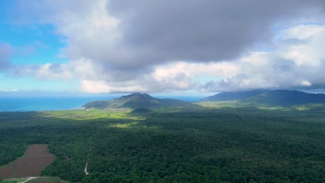 drone footage glides over dense rainforest toward distant mountains and coastline under dramatic clouds, with sunlight illuminating green canopy and agricultural clearings