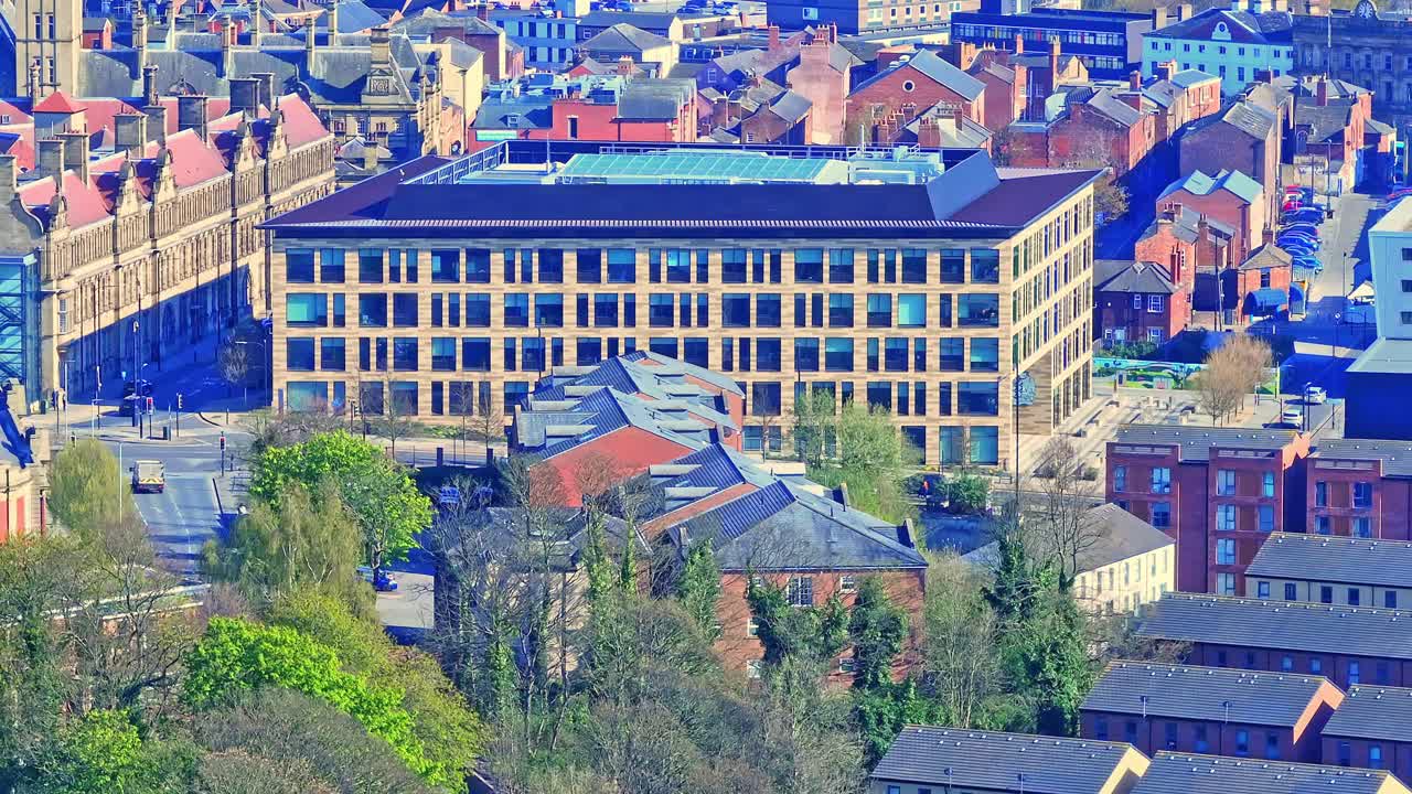 Aerial view of Wakefield city centre, Yorkshire, UK, in morning sunlight, featuring urban buildings, offices, moving traffic on roads, and spring trees adding vibrant greenery to the streetscape.