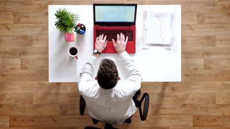 joven escribiendo en una computadora portátil y mirando la pantalla, topshot, sentado detrás de la mesa con documentos y café