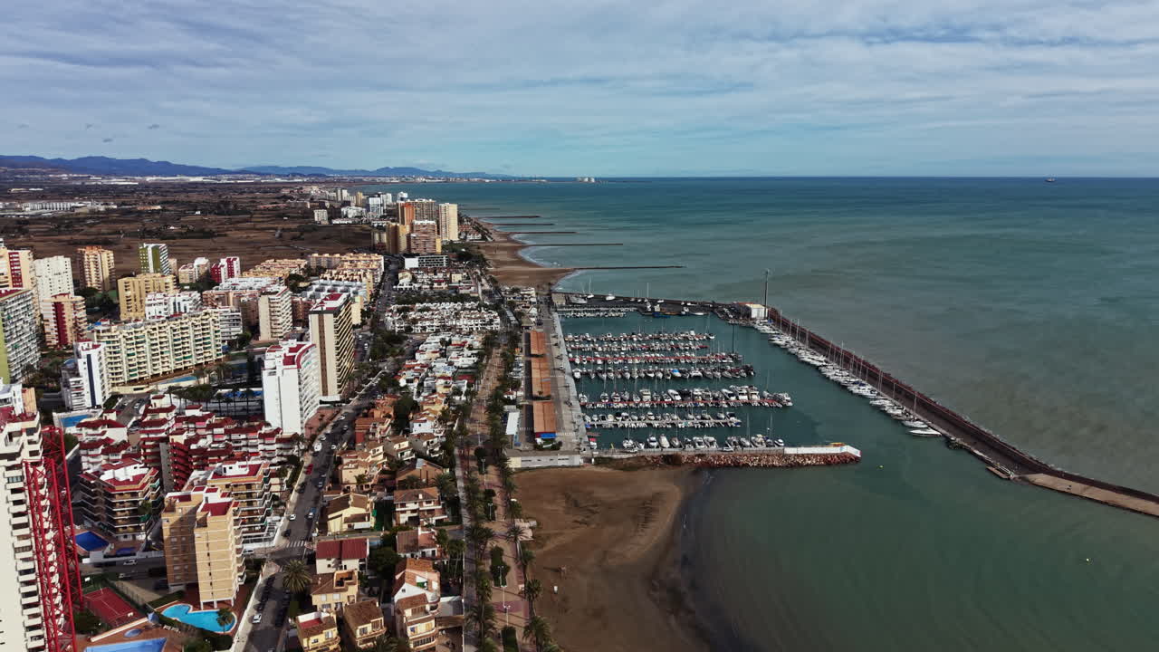 Kystudsigt over en marina med både, bygninger og strand under en overskyet himmel i Valencia, Spanien.