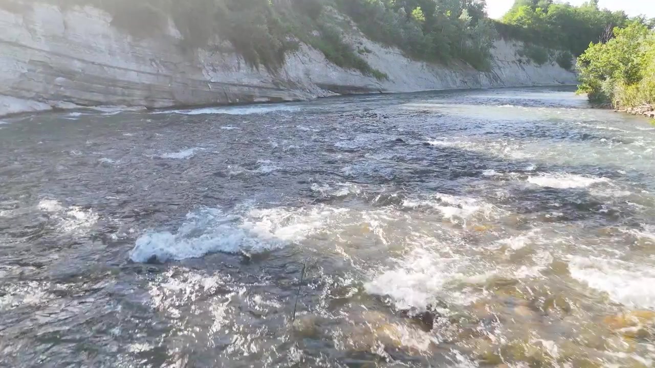 Inquadratura ampia statica di un fiume impetuoso accanto a ripide scogliere calcaree sotto la luminosa luce estiva.