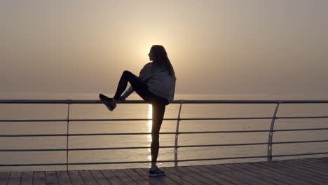 Slim-woman-with-long-legs-doing-stretching-on-sunrise-by-the-seaside,-perfect-body.-Stretches-her-leg-above-head.-Backside-view