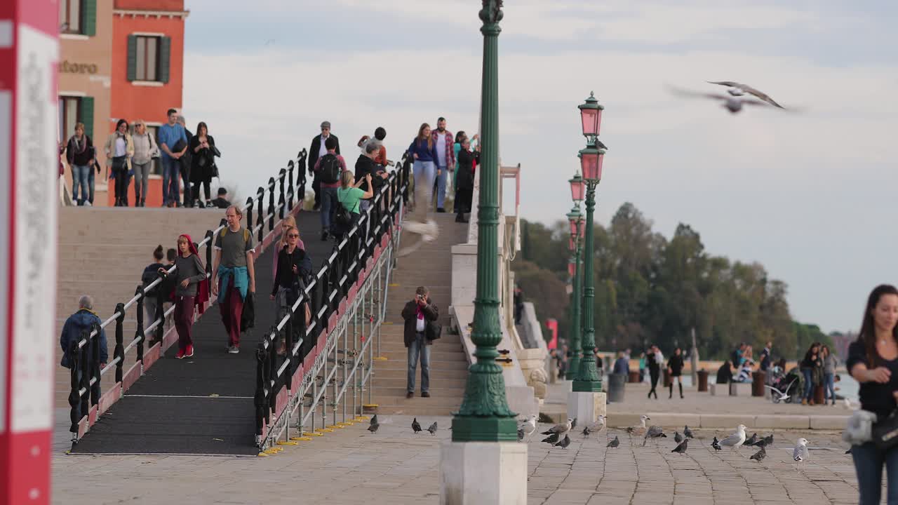 Tourists walking on historic grand canal main street passing bridge over the canal with beautiful old green metal lantern and flying birds