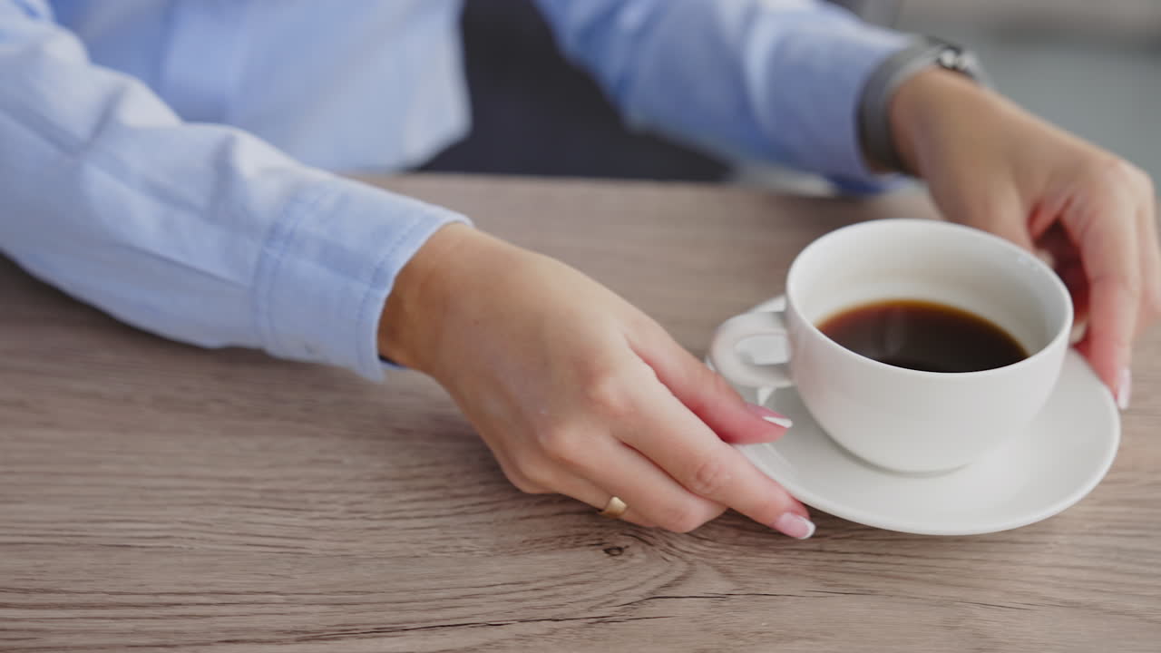 White cup of black coffee on the wooden table. Female hands pull the cup closer. Laptop and phone are on the desk beside. Close up.