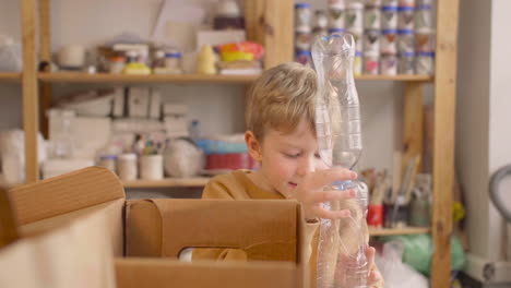 blond kid putting plastic bottles in cardboard box on a table in a craft workshop