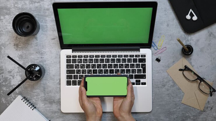 Woman works using several devices laptop and smartphone with green screen. Young creative female working on Mock-up laptop while sitting at office work desk. Modern workplace. Chroma key
