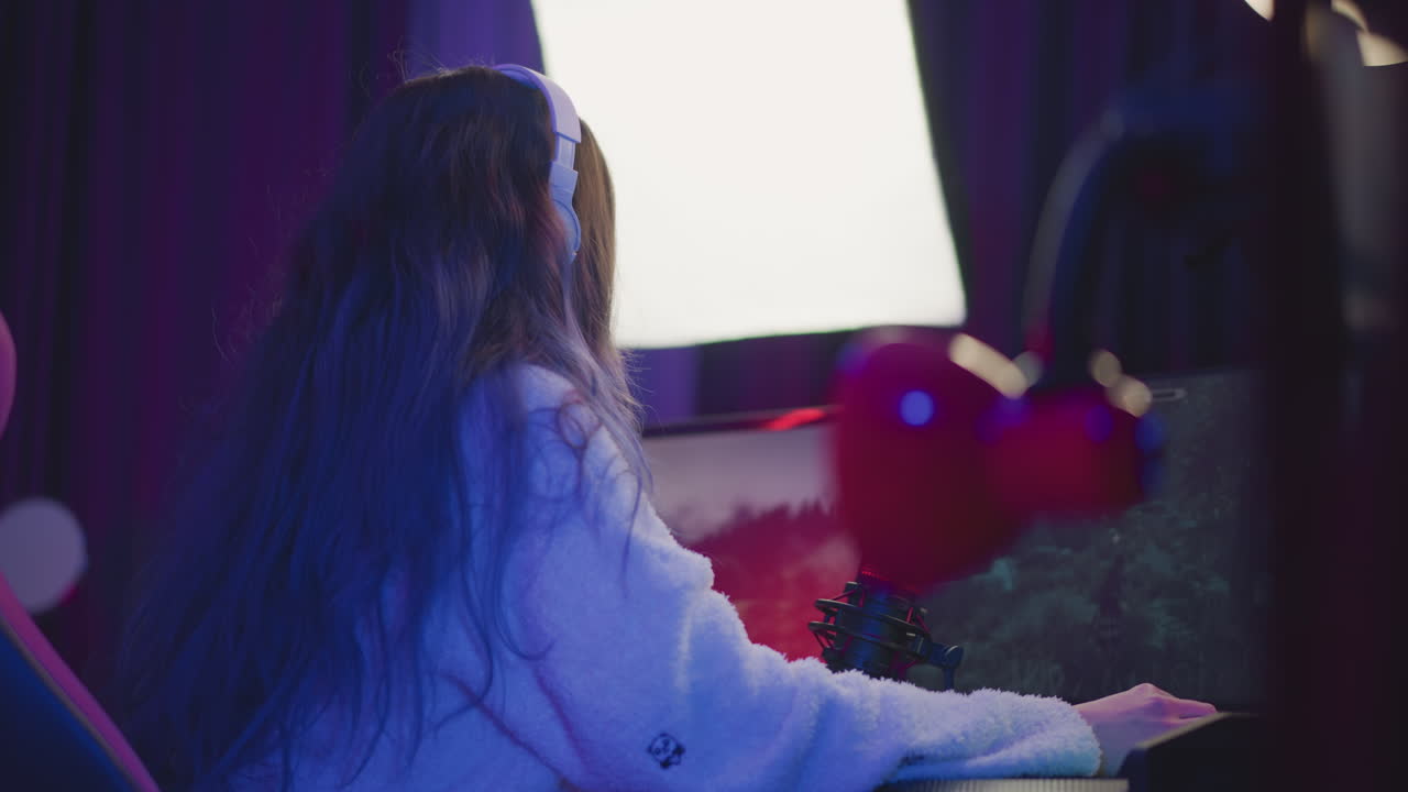 back view of woman with long wavy hair cascading down shoulders wearing white headset and fluffy sweater focused on playing immersive video game on dual monitors with red mic and ambient rgb lighting