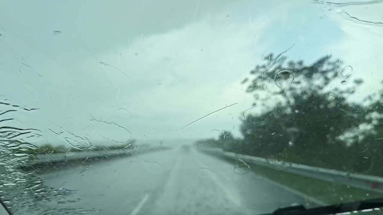 La pioggia cade su una strada mentre un veicolo avanza durante una giornata nuvolosa in una località sconosciuta.