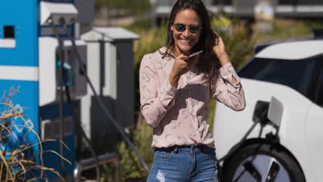 mujer caucásica sonriente usando un teléfono inteligente y cargando un coche eléctrico