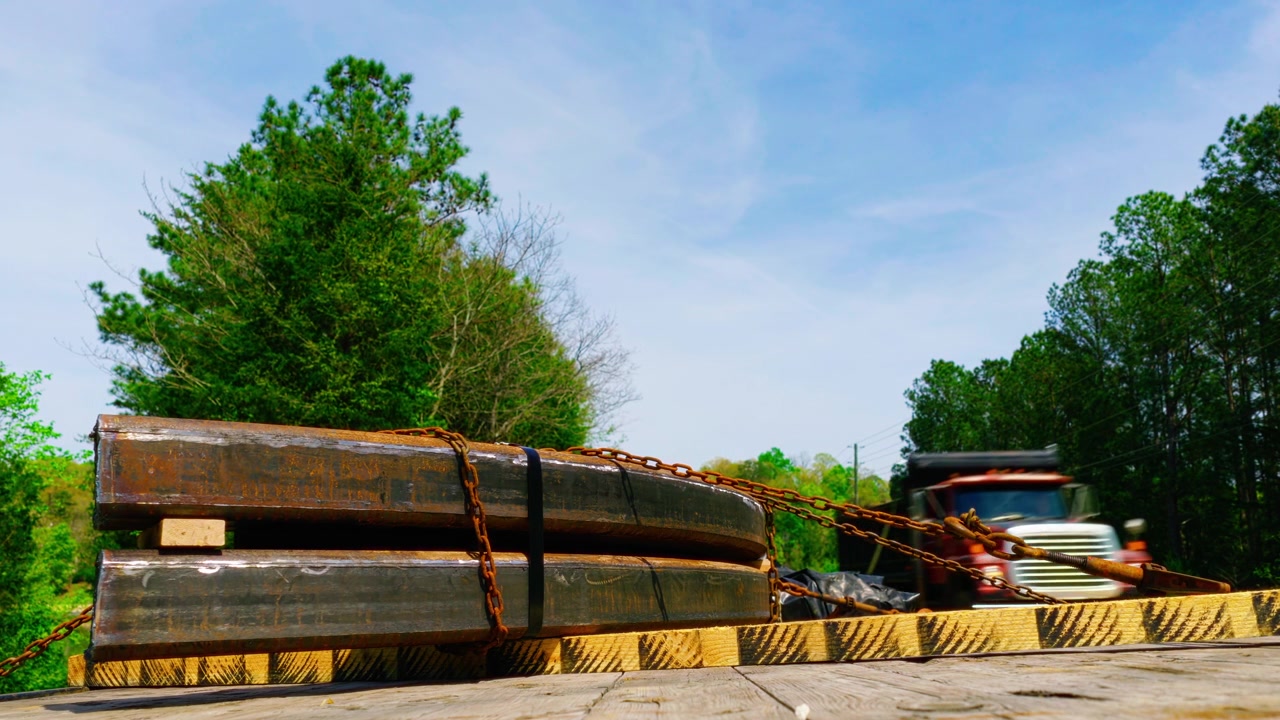 Industrial steel beams secured with binding chains and straps on a flatbed trailer, illustrating heavy cargo transport, raw material supply chains, and B2B freight distribution.