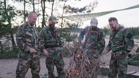 conversación de soldados en ropa de camuflaje, están discutiendo por la noche junto a la fogata común fritando salchichas para la cena, los hombres están descansando después del trabajo, servicio, ejercicios de campo