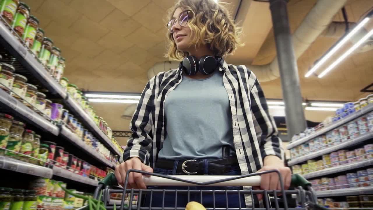 목에 카레드 셔츠와 헤드폰을 입은 쾌활한 여성이 식료품 가게에서 음식이 들어있는 쇼핑 트롤리를 운전하고 있습니다.