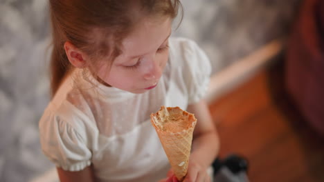 overhead view of young girl with light brown hair wearing white blouse holding ice cream cone, glancing thoughtfully at background wall while continuing to enjoy cone indoors on wooden floor setting