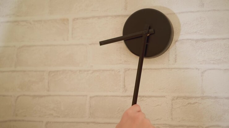 Woman with long hair and light brown clothing carefully adjusts hands of modern black wall clock mounted on white brick wall. scene is calm indoors with soft ambient daylight illuminating