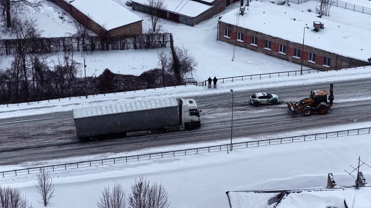 Aerial drone shot of a semi trailer on a slippery winter road as a wheel loader transports a stranded car, showing icy conditions, traffic risk, and recovery work