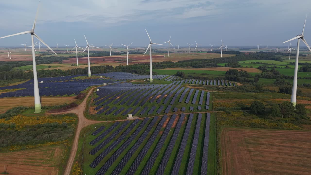 Aerial view of a renewable energy site featuring rows of solar panels and wind turbines spread across green agricultural fields under a partly cloudy sky