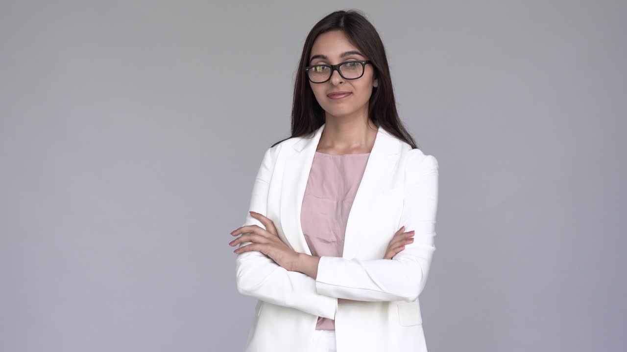 Retrato de una feliz, hermosa, joven y elegante mujer de negocios india, vistiendo traje blanco y gafas, mirando a la cámara y sonriendo con confianza. Esta bonita profesional hindú posa con los brazos cruzados sobre un fondo de estudio gris.