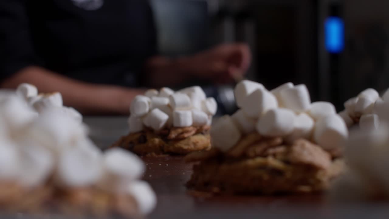 Pasticcere che tosta marshmallow con una torcia da cucina sui biscotti a un bancone di dolci, con messa a fuoco selettiva e luce calda