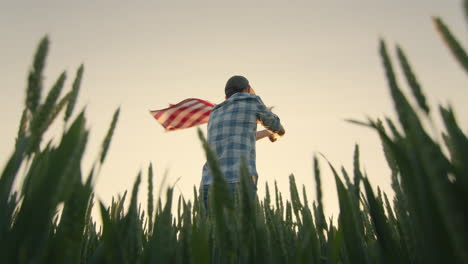 Silueta-De-Un-Joven-Ondeando-La-Bandera-Americana.-De-Pie-En-Un-Campo-De-Trigo-Al-Atardecer.-Tiro-Amplio-De-ángulo-Bajo