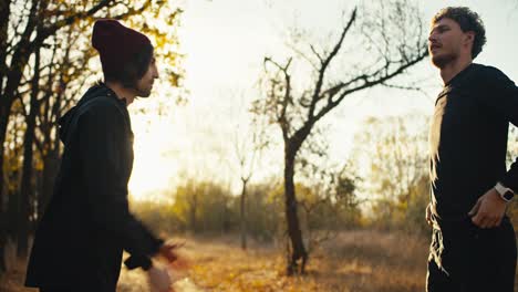 a happy brunette man in a red hat and a black sports summer uniform together with his friend a man with curly hair warms up before their jog and then high-fives each other and begins their run along an earthen path in the autumn summer forest