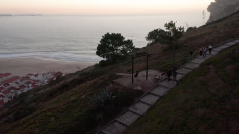 Tourists-On-Walking-Trail-Of-Hill-Overlooking-Town-And-Beach-Of-Nazare-In-Portugal---aerial-drone-shot