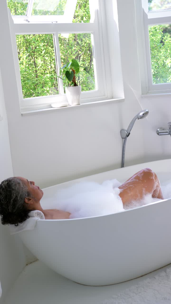 Donna afroamericana sdraiata in una vasca da bagno a libera installazione con schiuma in un bagno bianco