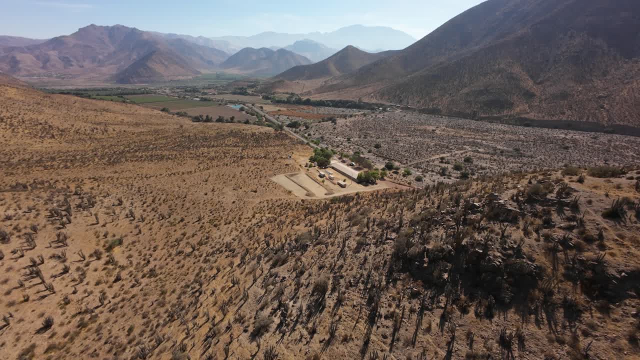Cinematic drone shot over an arid valley with scattered vegetation, farmlands, and rugged mountains under a clear sky. Ideal for nature, travel, or documentary content