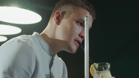 close-up side shot of player in white shirt holding cue stick, sipping lemon drink with black straw. overhead lamp light highlights focused expression in moody pool hall atmosphere