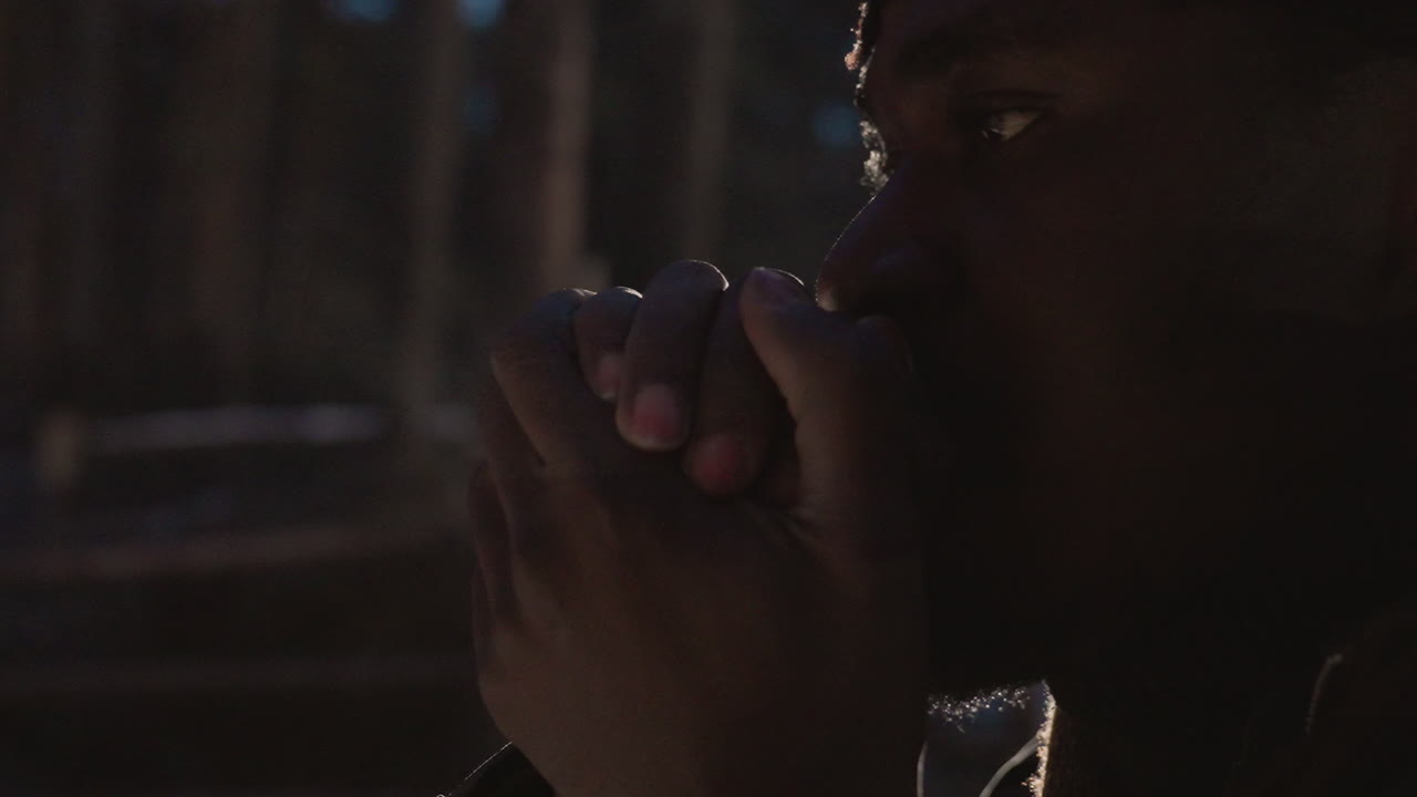 hombre negro creando una llama con una cerilla, con las manos ahuecadas protegiendo la chispa, brillo cálido en la cara, luz de la brasa contra el bosque oscuro, expresión de concentración, acción de supervivencia primitiva, textura cinematográfica íntima