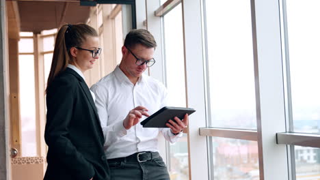colleagues working in office stand at the window. man uses i-pad and woman points at something on gadget.