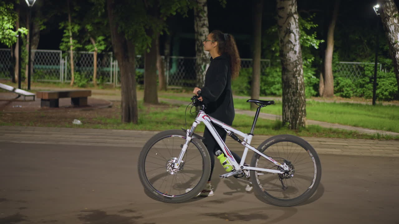 Female Cycling Evening, Woman Riding Bicycle In Park, Woman Cycling Leisurely Through Park With Soft Lamplight Glow, Woman Leisurely Rides Her Bicycle Along Park Pathway In Quiet Evening
