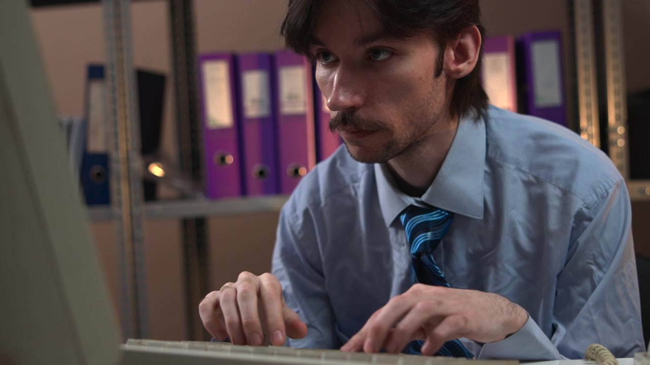 A focused young man in a light blue shirt and striped tie works on a laptop while seated at a desk in a quiet office, with purple binders and shelving visible