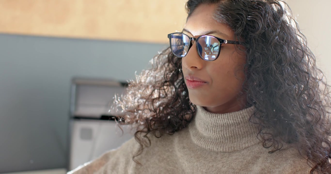 Donna afroamericana in ufficio, che sente una voce, si sistema gli occhiali e il dolcevita mentre legge il monitor.