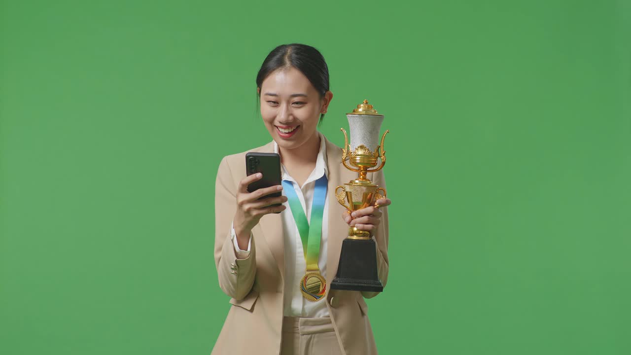 골드 메달을 들고 금 트로피를 들고 스튜디오의 녹색 화면 배경에 스마트폰을 사용하는 아시아 사업가 여성