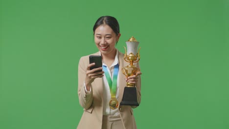 골드 메달을 들고 금 트로피를 들고 스튜디오의 녹색 화면 배경에 스마트폰을 사용하는 아시아 사업가 여성