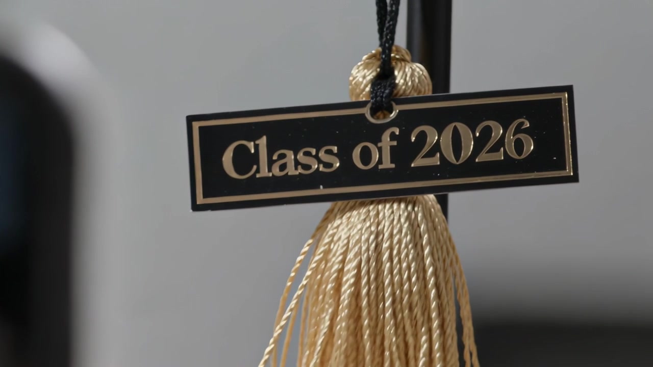 Hände, die ein Telefon halten, bewegen sich näher zu einer goldenen Quaste in einem Schulbereich und enthüllen die Aufschrift „Class of 2026“.