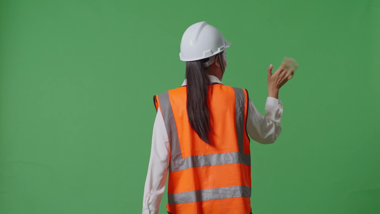 안전 헬을 입은 아시아 여성 엔지니어가 녹색 스크린 배경 스튜디오에서 는 동안 손을 흔들고 있는 뒷면