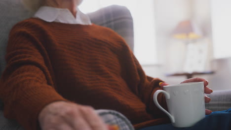 senior woman with hot drink keeping warm by portable radiator at home in cost of living crisis