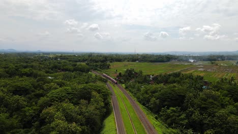 drone view following train crossing bridge in tropical countryside, indonesia