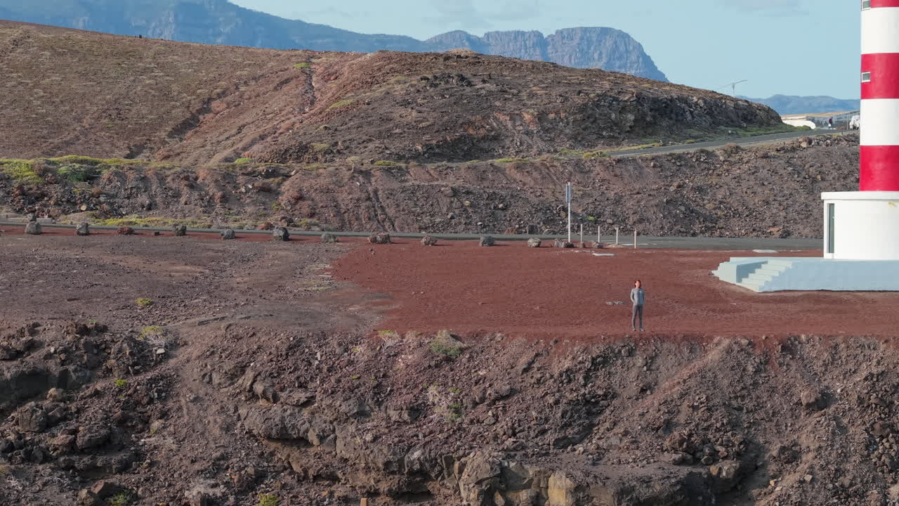 Zooming out aerial view of a woman walking near the Punta Sardina lighthouse on the rocky coast of Gran Canaria