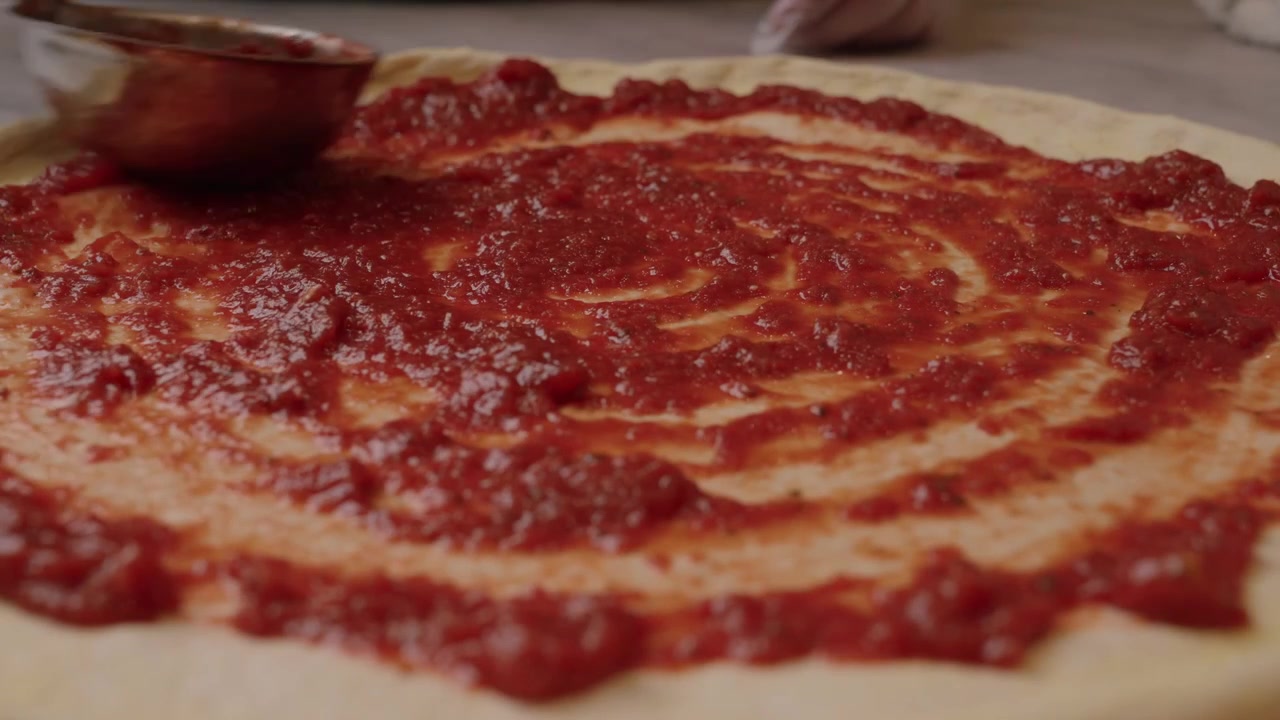 Un mestolo di metallo spalma una ricca salsa di pomodoro rossa su impasto per pizza appena preparato in una scena di cucina ravvicinata.
