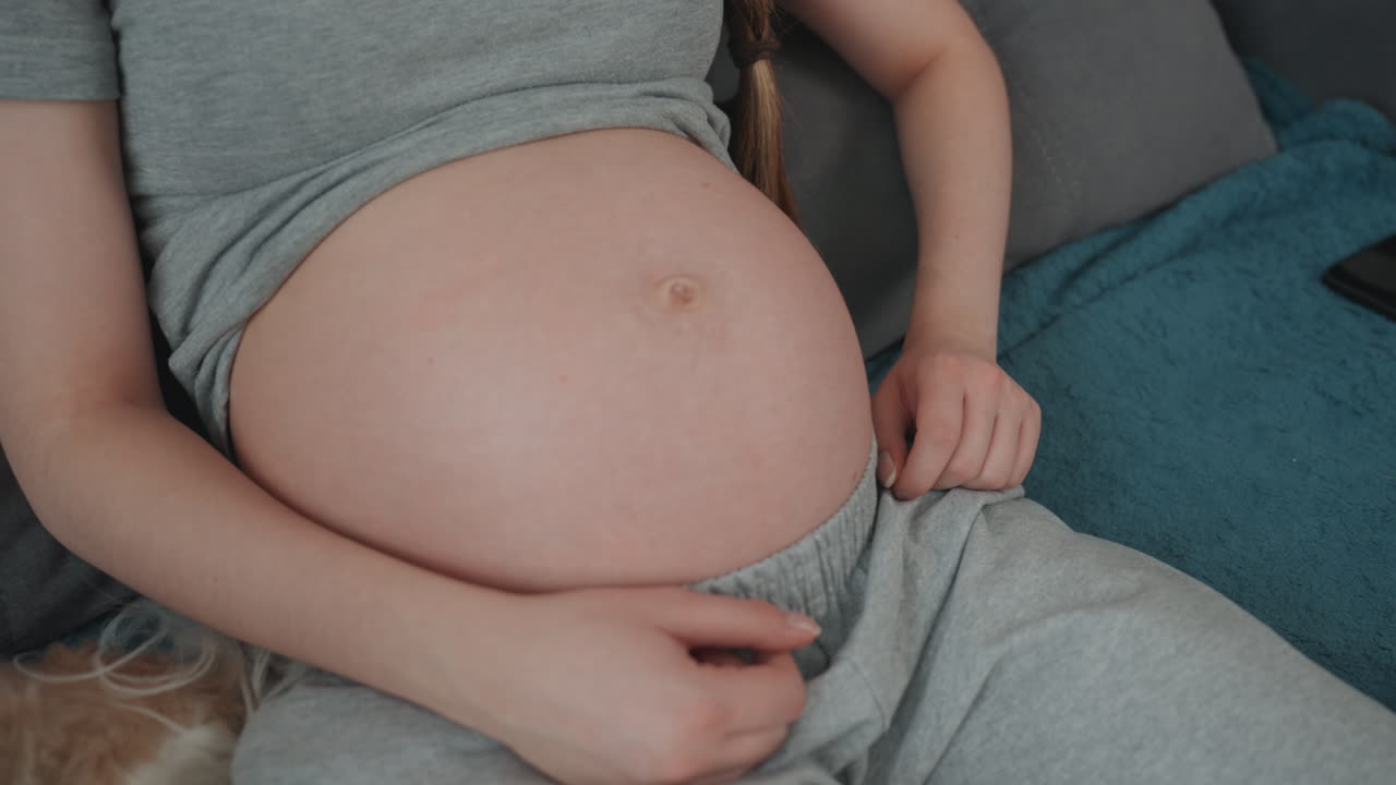 Pregnant white woman examining belly closely, fingers near navel counting kicks, focused expression, braided hair, loungewear, closeup living room shot, natural light, mindful prenatal check