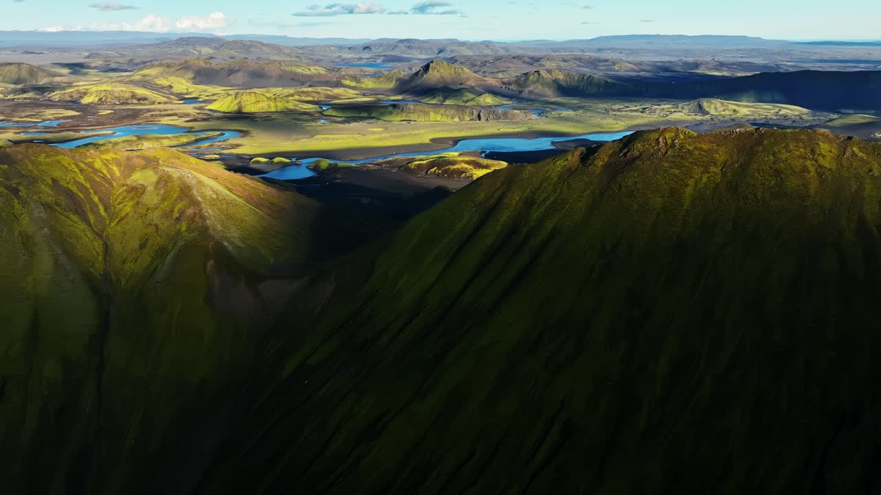 Sunlit Icelandic highlands reveal bright moss ridges and winding blue rivers across a vast volcanic plateau, captured from above in golden-hour light