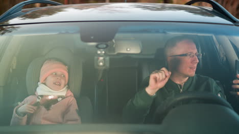 a joyful in a car, showing a man holding a smartphone while seated in the driver's seat, with a young girl beside him in the front passenger seat. both are smiling and appear to be dancing together