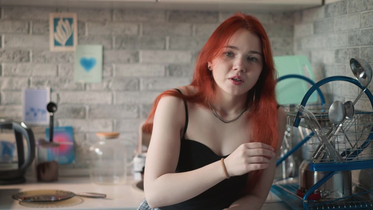 Donna dai capelli rossi che si protende sul bancone, cucina moderna con paraschizzi in mattoni, maglietta nera casual, che maneggia un dolce mentre riflette sulla prossima mossa, luce soffusa dalla finestra, quiete domestica, sottile prescienza.