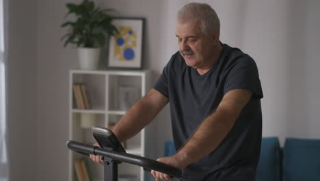 Un-Hombre-De-Mediana-Edad-Y-Cabello-Gris-Está-Entrenando-En-Bicicleta-Estática-En-Casa-Preocupándose-Por-La-Salud-Y-Manteniéndose-En-Forma-En-La-Actividad-Deportiva-De-La-Vejez