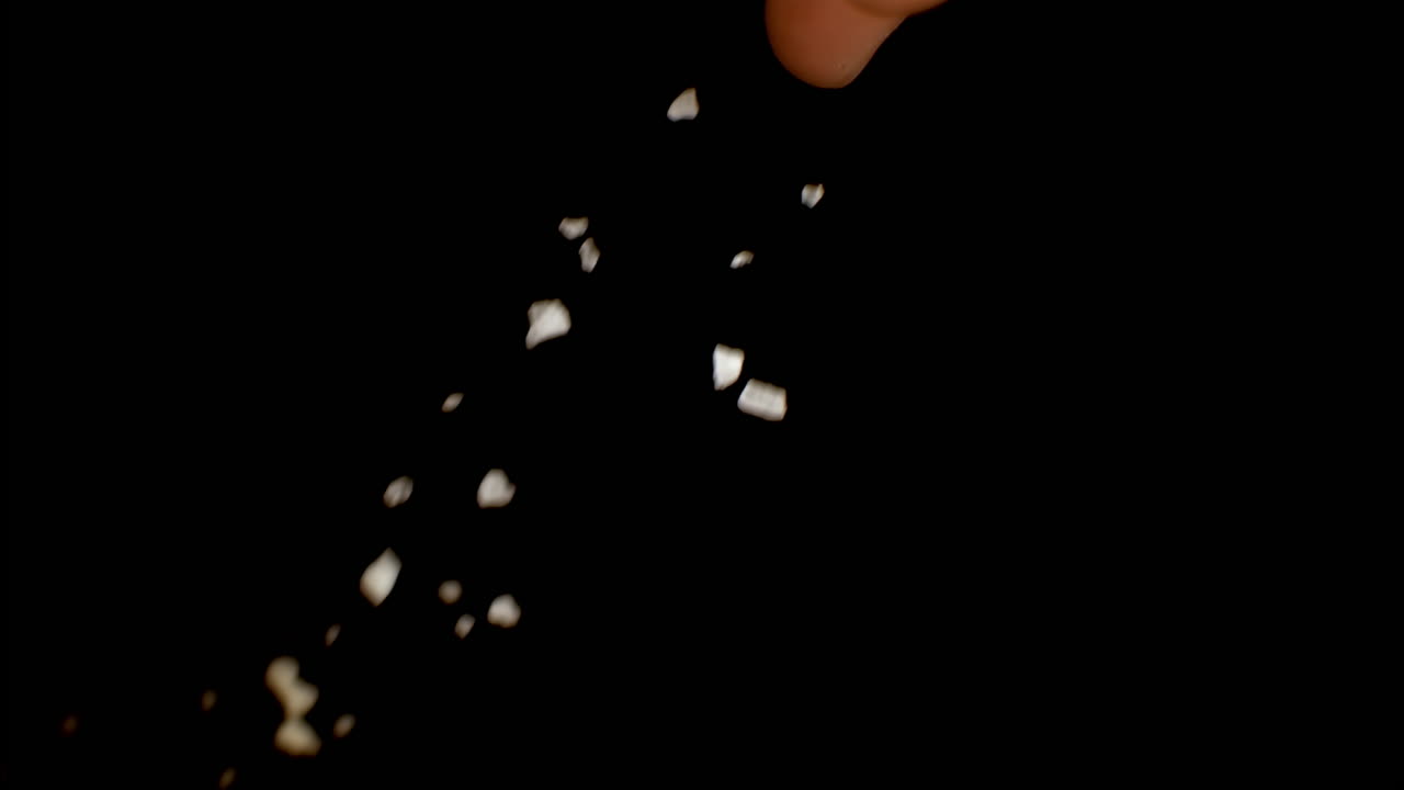 Coarse salt falling in slow motion on black background, macro food detail handheld