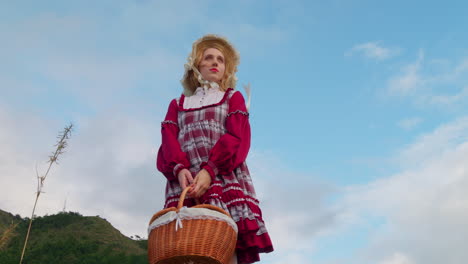 vista de bajo ángulo de modelo caucásico rubio en vestido renacentista medieval tradicional de moda antigua sosteniendo una canasta de alimentos