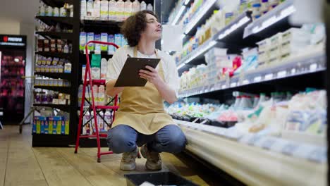 A-guy-with-curly-hair-with-a-tablet-in-his-hands-takes-inventory-of-goods-in-the-dairy-products-compartment-in-a-supermarket