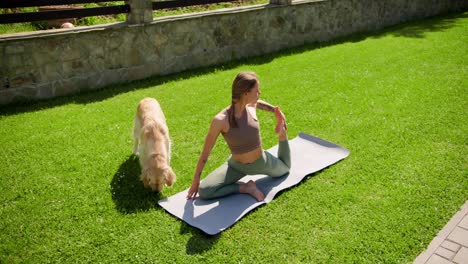 A-blonde-girl-in-a-special-sports-summer-uniform-does-yoga-on-a-gray-rug-on-the-lawn,-her-light-colored-dog-walks-beside-her